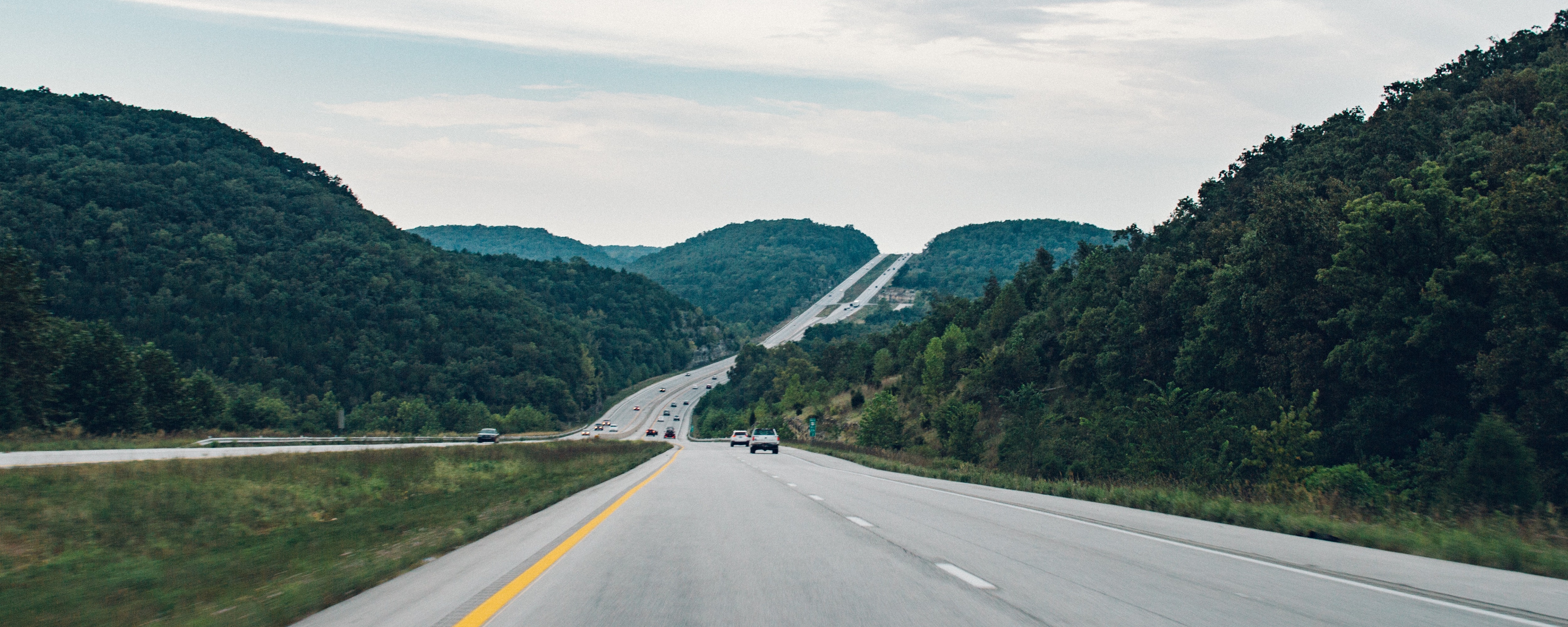Highway with several cars extends past rolling forested hills.